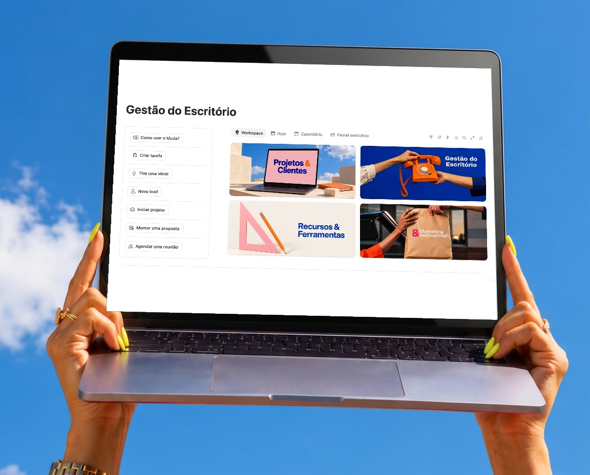 Notebook segurado pelas mãos contra o céu azul, com a tela oficial do sistema Muda